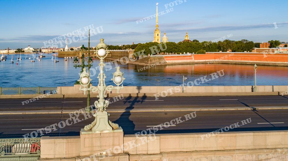 На этой фотографии солнечный свет мягко освещает Неву и Троицкий мост в Санкт-Петербурге. Золотые шпили Петропавловской крепости возвышаются на фоне, добавляя историческую значимость пейзажу. Это изображение подходит для туристических публикаций, утренних тем в средствах массовой информации и для тех, кто хочет украсить свое пространство видом на величественный Петербург.